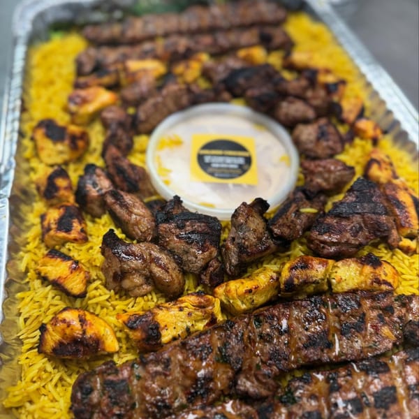 A catering tray filled with yellow rice, grilled chicken, beef kebabs, and a sauce container.